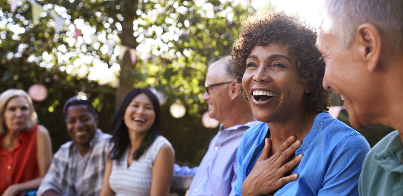 A diverse group of people, including both men and women of different ages, enjoying a lively conversation outdoors in a garden setting. They are smiling and laughing, creating a warm and joyful atmosphere, with festive decorations hanging in the background.